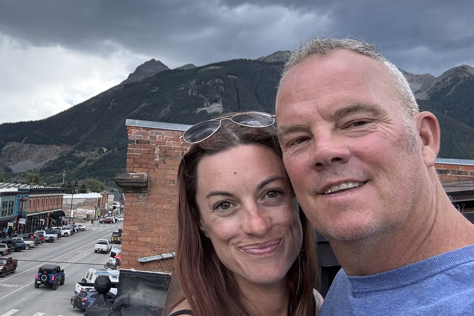Holland Duell and Alyssa Arroyo smiling close together in a selfie on a rooftop in Silverton, Colorado, with the town's brick main street and forested mountains under a stormy sky behind them. Alyssa has sunglasses pushed up in her hair; Holland wears a blue t-shirt.